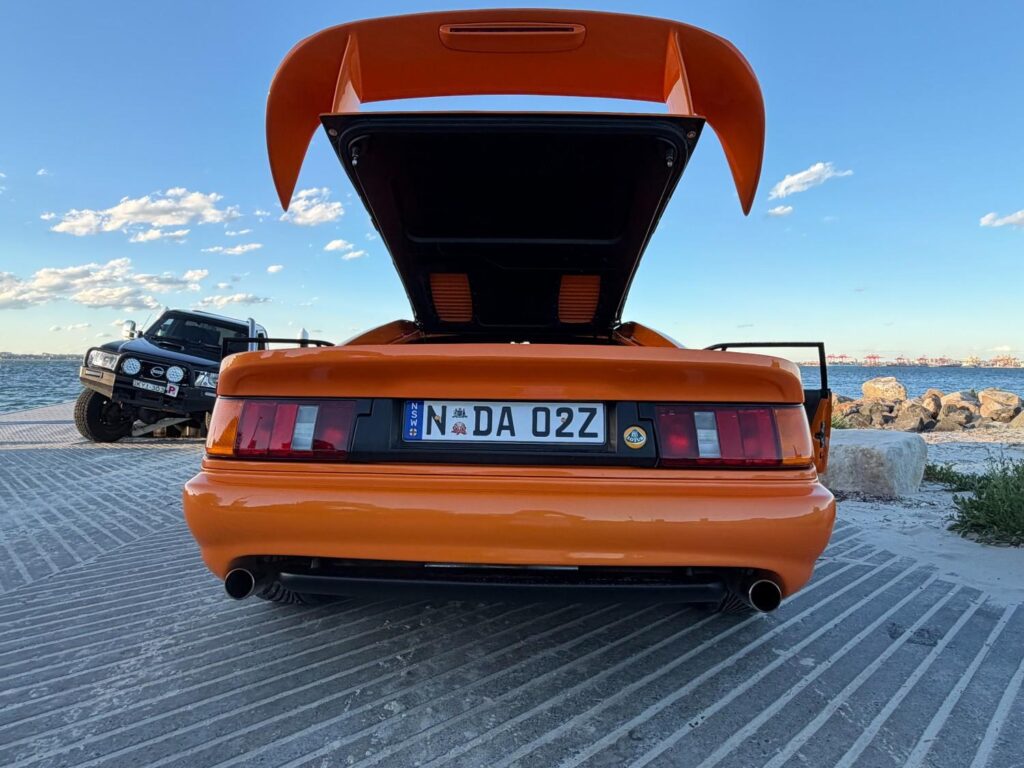 Orange 1999 Lotus Esprit GT3 wide rear view with the hatch raised on a boat ramp by the sea